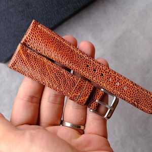 Puede incluir: Una correa de reloj de color tostado con una superficie texturizada y granulada. La correa cuenta con una hebilla de metal plateado y una serie de orificios de ajuste a lo largo de la banda. El material tiene un grano distintivo y un tono anaranjado.