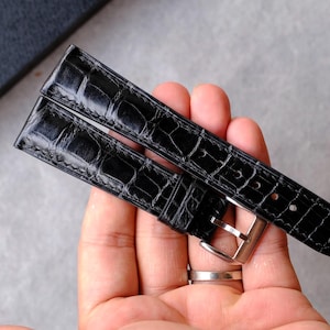 Puede incluir: Correas de reloj negras con textura de piel de cocodrilo. Las correas tienen una hebilla de color plateado y se sostienen en una mano. Las correas de reloj son nuevas.