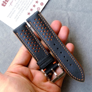 Puede incluir: Una correa de reloj de cuero negro con un diseño perforado, una parte inferior de color naranja brillante y costuras de contraste en naranja. La correa incluye una hebilla de metal plateado y dos trabillas de cuero con acabado texturizado.
