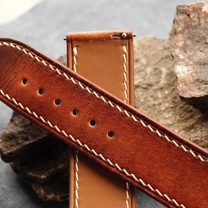 Pode incluir: Uma bracelete de rel&oacute;gio em pele castanha com um acabamento texturado e desgastado na superf&iacute;cie exterior e um verso liso de cor bege claro. A bracelete apresenta costuras grossas em contraste de cor creme nas bordas e inclui pinos met&aacute;licos.