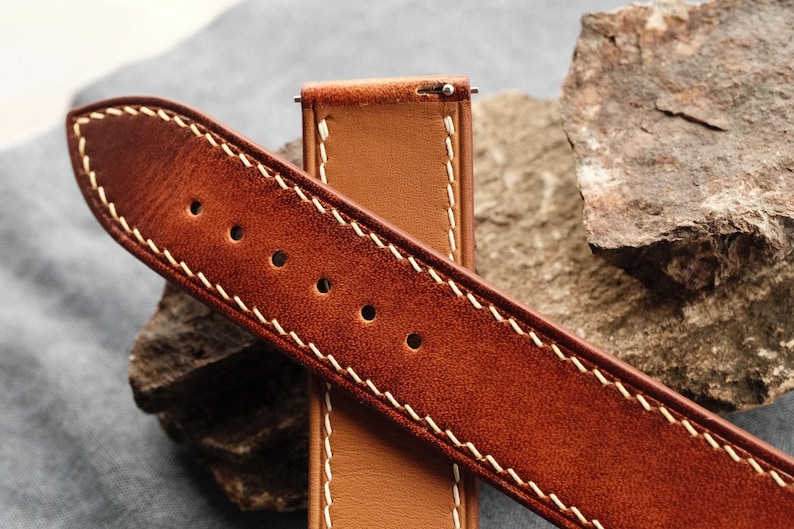 Pode incluir: Uma bracelete de rel&oacute;gio em pele castanha com um acabamento texturado e desgastado na superf&iacute;cie exterior e um verso liso de cor bege claro. A bracelete apresenta costuras grossas em contraste de cor creme nas bordas e inclui pinos met&aacute;licos.