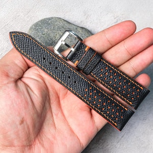 Op de afbeelding: Een zwarte lederen horlogeband met een getextureerd oppervlak, perforatiegaatjes en feloranje stiksels langs de randen. De band is voorzien van een gepolijste zilverkleurige metalen gesp en twee bijpassende lederen lussen voor bevestiging.