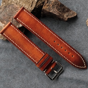 Pode incluir: Uma bracelete de rel&oacute;gio de duas pe&ccedil;as feita de couro liso cor de conhaque com um acabamento degrad&eacute;. A bracelete inclui costuras brancas em contraste ao longo das bordas, dois passadores de couro e uma fivela de metal prateado polido.