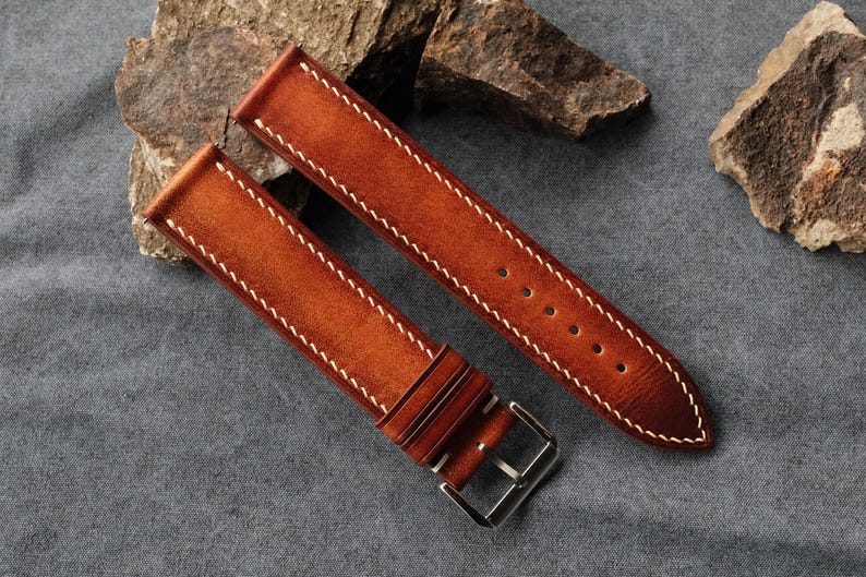 Pode incluir: Uma bracelete de rel&oacute;gio de duas pe&ccedil;as feita de couro liso cor de conhaque com um acabamento degrad&eacute;. A bracelete inclui costuras brancas em contraste ao longo das bordas, dois passadores de couro e uma fivela de metal prateado polido.