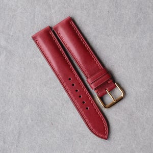 May include: A deep red leather watch strap featuring tonal stitching along the edges, a series of six circular adjustment holes, and a polished gold-tone metal pin buckle. The strap consists of two separate pieces laid side by side.