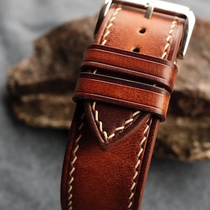 Pode incluir: Uma pulseira de rel&oacute;gio em couro castanho com costuras vis&iacute;veis em cor creme ao longo das bordas, dois passadores de couro e uma fivela de metal prateado polido. O couro tem um acabamento liso e ligeiramente polido com tons castanhos.