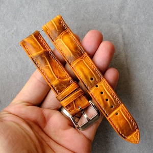 May include: A pair of watch straps in golden-brown leather with a crocodile-like texture. The straps have a silver buckle and are new. The watch straps are designed to be attached to a watch.