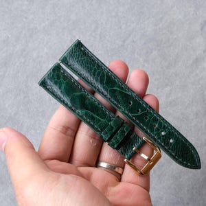 Op de afbeelding: Een donkergroene horlogeband met een gestructureerd, struisvogelachtig lederpatroon en zichtbare stiksels langs de randen. De band is voorzien van een gepolijste goudkleurige metalen gesp en twee lederen lussen voor bevestiging aan een horloge.