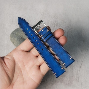 May include: A blue leather watch strap featuring a gradient finish, light-colored stitching along the edges, and a silver-toned metal buckle. The strap has a series of punched holes for adjustment and a smooth, polished surface texture.