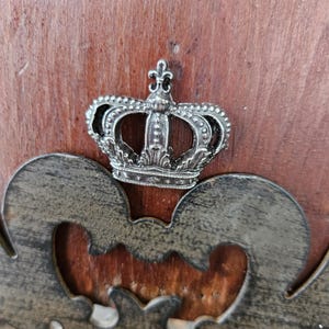 Pode incluir: Uma coroa prateada detalhada, com uma flor-de-lis, sobre um fundo de madeira escura. A coroa é o elemento central, com um design decorativo de metal por baixo.