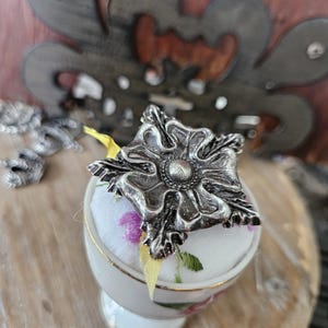 Pode incluir: Um alfinete de flor prateado e ornamentado repousa sobre uma pequena almofada de alfinetes com padrão floral. A almofada de alfinetes está colocada num suporte decorativo branco em forma de ovo com uma borda dourada. Uma fita amarela é visível.