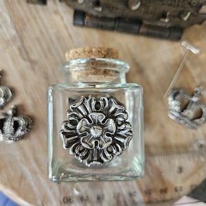 Puede incluir: Una botella cuadrada de vidrio transparente con tapón de corcho y adorno floral plateado. La botella está decorada con un diseño floral plateado. Artículos decorativos.