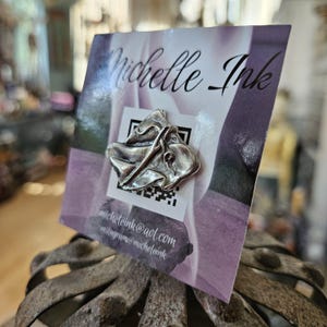 May include: A silver-toned metal pin in an abstract design, attached to a card with the text "Michelle Ink". The card has a QR code and contact information. The background is a soft purple and white floral pattern.
