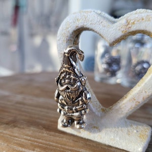 Könnte beinhalten: Ein silberner Anhänger in Form eines Zwergs mit Bart und spitzem Hut. Der Anhänger liegt auf einer weißen herzförmigen Oberfläche.