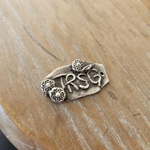 Könnte beinhalten: Ein silberfarbener Metallanhänger mit den Buchstaben "RSG" und drei floralen Verzierungen. Der Anhänger hat eine strukturierte Oberfläche und ein kleines Loch oben. Der Anhänger liegt auf einer hellbraunen Holzoberfläche.