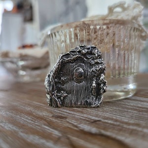 Könnte beinhalten: Ein silberner Anhänger in Form einer kleinen Tür mit einem runden Fenster, umgeben von Ranken und Blättern.