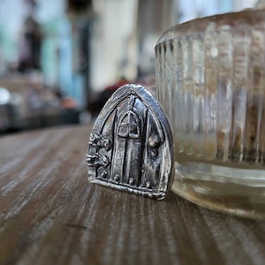 Könnte beinhalten: Ein silberner Anhänger in Form einer kleinen, verzierten Tür mit einem detaillierten Design. Die Tür hat ein kleines Fenster und ist von dekorativen Elementen umgeben.