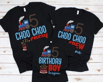 May include: Black t-shirts with train and number 5 graphics. The shirts read "CHOO CHOO CREW" and "BIRTHDAY BOY" with a name. A birthday cake with candles is also featured. The shirts are for a birthday celebration.