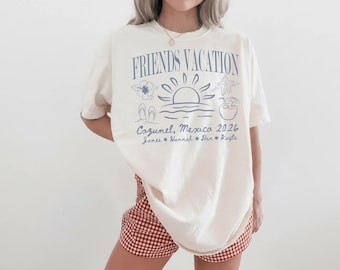 May include: Cream-colored t-shirt with the text "FRIENDS VACATION" and a beach-themed graphic in blue. The graphic includes a sun, waves, and tropical elements. Below, it reads "Cozumel, Mexico 2026" and names. The person is wearing red and white checkered shorts.