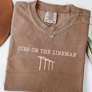 May include: A brown Comfort Colors t-shirt with the text "DIBS ON THE LINEMAN" and a graphic of power lines. A silver necklace with a small pendant rests on the shirt. A brown hat and dried plant are in the background.