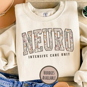 May include: Cream-colored sweatshirt with the word "NEURO" in a floral design, above "INTENSIVE CARE UNIT" in black text. A small, round graphic says "HOODIES AVAILABLE". A hat and jeans are in the background.
