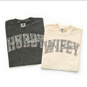 May include: Two t-shirts, one dark gray and one cream-colored, are displayed. The dark gray shirt says "Hubby Mr. Smith" in a camouflage-style font. The cream shirt says "Wifey Mrs. Smith" in a similar font. Both shirts are folded.