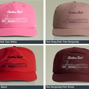 May include: Four different colored baseball caps with a surfboard graphic and the text "Custom Text" on the front. The caps are in the colors charity pink, hazy pink, red, and burgundy. The text on the caps is in the colors white, burgundy, black, and storm.
