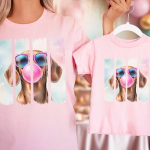 May include: Pink t-shirts with a graphic of a dachshund wearing sunglasses and blowing a bubble. The dog is in front of a colorful background.