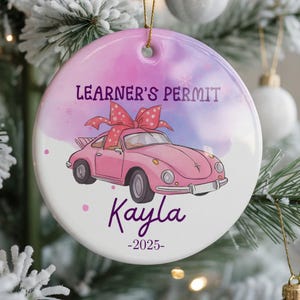 May include: A round, white ceramic ornament with a pink and purple watercolor background. The ornament features a pink car with a bow, the text "LEARNER'S PERMIT", the name "Kayla", and the year "-2025-". A gold string is attached for hanging.