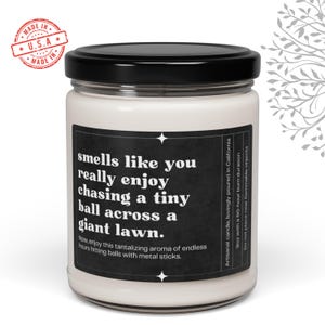 May include: A white candle with a black label that reads "smells like you really enjoy chasing a tiny ball across a giant lawn. Now, enjoy this tantalizing aroma of endless hours hitting balls with metal sticks." The label also includes the text "Artisanal candle, lovingly poured in California 9oz with a 50-hour burn duration. Do not place near flammable objects."