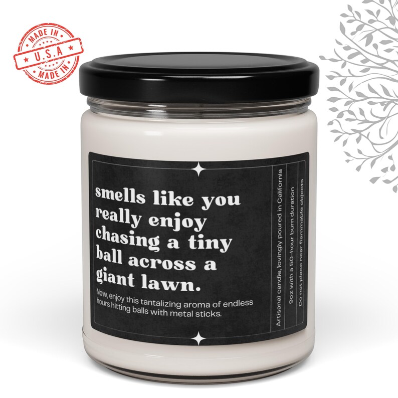 May include: A white candle with a black label that reads "smells like you really enjoy chasing a tiny ball across a giant lawn. Now, enjoy this tantalizing aroma of endless hours hitting balls with metal sticks." The label also includes the text "Artisanal candle, lovingly poured in California 9oz with a 50-hour burn duration. Do not place near flammable objects."