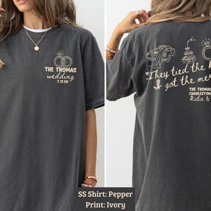 May include: Dark gray t-shirt with ivory print. Front features wedding rings and text "THE THOMAS wedding 7.12.26". Back has text "They tied the knot. I got the merch." with wedding-themed graphics and names.
