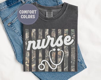 Pode incluir: Camiseta cinza escuro com a palavra "nurse" em branco e um gráfico de estetoscópio branco. A camisa tem um padrão de camuflagem atrás da palavra "nurse" e é exibida em jeans azul.