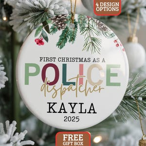 May include: A white ceramic Christmas ornament with the text "FIRST CHRISTMAS AS A POLICE dispatcher KAYLA 2025". The ornament has a gold string for hanging and is decorated with green and red floral accents. The words "4 DESIGN OPTIONS" and "FREE GIFT BOX" are also visible.