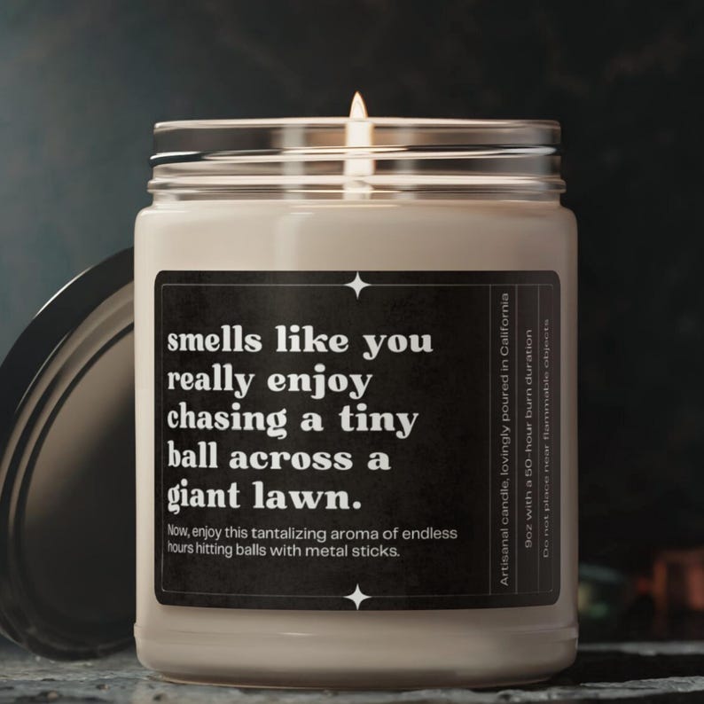 May include: A white candle in a glass jar with a black label that reads "smells like you really enjoy chasing a tiny ball across a giant lawn. Now, enjoy this tantalizing aroma of endless hours hitting balls with metal sticks." The label also includes the text "Artisanal candle, lovingly poured in California 9oz with a 50-hour burn duration Do not place near flammable objects."