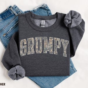 May include: A dark heather gray crewneck sweatshirt featuring the word GRUMPY in a collegiate font with a camouflage pattern fill. The sweatshirt is layered over a pair of blue denim jeans. The text DARK HEATHER is printed in the bottom corner.
