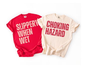 May include: Two t-shirts, one red and one cream-colored, are displayed. The red shirt has the text "SLIPPERY WHEN WET" in a distressed font. The cream shirt has the text "CHOKING HAZARD" in a similar font.