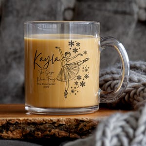 May include: A clear glass mug filled with a light brown liquid, featuring a black illustration of a ballerina and snowflakes. The mug has the name "Kayla" and text related to "The Nutcracker" ballet.