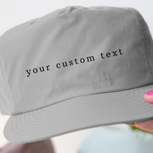 May include: A light gray baseball cap with a curved brim. The text "your custom text" is printed across the front in a black, sans-serif font. The cap is held by a person with pink nail polish.