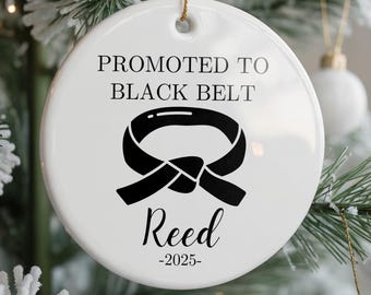 May include: A white ceramic Christmas ornament with the text "PROMOTED TO BLACK BELT" and a black belt graphic. Below the graphic is the name "Reed" and the year "-2025". The ornament is hanging from a gold string.