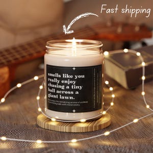 May include: A lit candle in a clear glass jar with a black label that reads "smells like you really enjoy chasing a tiny ball across a giant lawn. Now enjoy this tantalizing aroma of endless hours hitting balls with metal sticks."