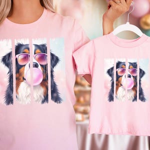 May include: Pink t-shirts with a graphic of a Bernese Mountain Dog wearing sunglasses and blowing a bubble. The dog is in a colorful, abstract background.