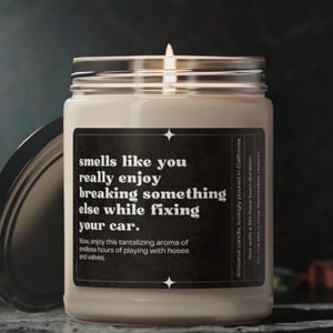 May include: A white candle in a glass jar with a black label that reads "smells like you really enjoy breaking something else while fixing your car. Now, enjoy this tantalizing aroma of endless hours of playing with hoses and valves." The label also includes the text "Artisanal candle, lovingly poured in California 9oz with a 50-hour burn duration. Do not place near flammable objects."