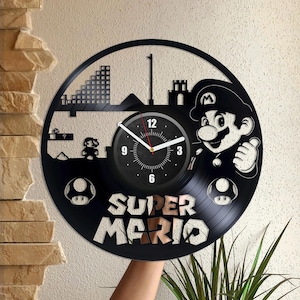 Könnte beinhalten: Schwarze Schallplattenuhr mit Super Mario-Motiv. Das Zifferblatt ist eine Schwarzweißillustration von Mario, einem Fragezeichenblock und einer Mauer aus Ziegeln. Die Zeiger der Uhr sind rot und weiß. Die Worte "Super Mario" sind in Gold auf die Schallplatte gedruckt.