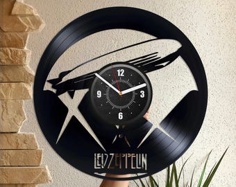 Könnte beinhalten: Eine schwarze Schallplattenuhr mit einem weißen Zifferblatt und schwarzen Zeigern. Die Uhr ist in Form eines Zeppelins gestaltet und trägt den Schriftzug "LED ZEPPELIN".