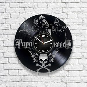 Puede incluir: Reloj de vinilo negro con una imagen de banda y el texto "Papa Roach". La esfera del reloj tiene un diseño de calavera y huesos cruzados con alas.