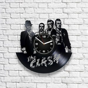 Op de afbeelding: Een zwarte vinyl platenspeler klok met een silhouet van de band The Clash. De wijzerplaat van de klok is wit met zwarte cijfers en wijzers. De naam van de band is in witte letters op de plaat geschreven.