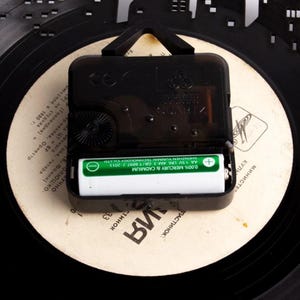 May include: Close-up of a black clock mechanism with a silver battery. The clock mechanism is labeled "QUARTZ" and has a CE mark. The battery is labeled "0.00% MERCURY & CADMIUM AA 1.5V LR6 AM-3 GB/T 8897.2-2013 SHENZHEN YUNMAI TECHNOLOGY CO.LTD".