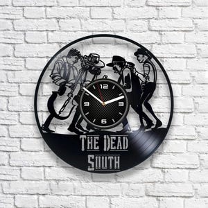 Puede incluir: Reloj de vinilo negro con una silueta de los miembros de la banda The Dead South. La esfera del reloj es blanca con números y manecillas negros. El nombre de la banda está impreso debajo de la esfera del reloj.