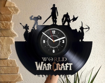 Puede incluir: Un reloj de vinilo negro con una silueta de personajes del videojuego World of Warcraft. La esfera del reloj es negra con números y manecillas blancas. El texto "World of Warcraft" está impreso en dorado en el disco.